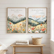 Two framed vintage Christian mountain landscape wall art prints featuring 2 Corinthians 12:9 and Isaiah 6:3 Scripture verses with wildflowers and evergreen forests, displayed side-by-side above a wooden bench in a bright, modern entryway.