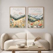 Two framed Christian mountain landscape wall art prints featuring 2 Corinthians 12:9 and Isaiah 6:3 Scripture verses with wildflowers and evergreen forests, displayed side-by-side above a cream bouclé sofa in a soft organic modern living room.