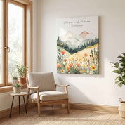 Unframed canvas Christian wall art featuring 2 Corinthians 12:9 Scripture verse with a watercolor mountain landscape and wildflowers, displayed on a light wall next to a wooden window and armchair in a sunlit room.