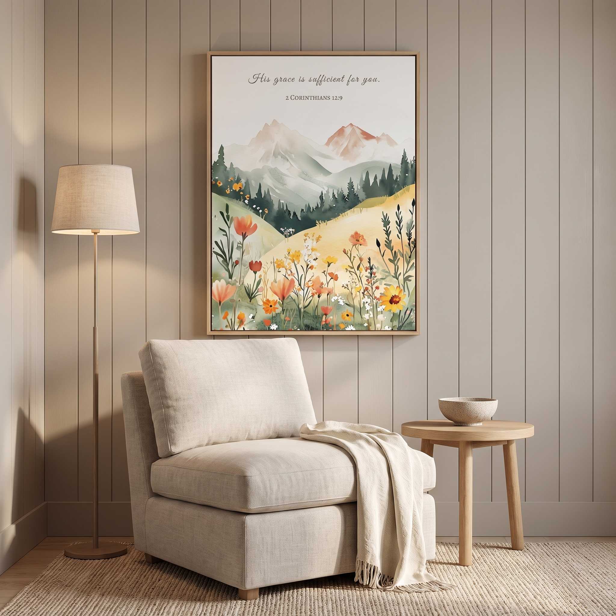 Framed Christian mountain landscape wall art titled 2 Corinthians 12:9 featuring mountains, pine trees, and wildflowers, displayed on a beige paneled wall above a neutral armchair in a cozy organic modern reading nook.