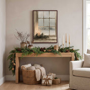 Framed Christmas candle and winter window wall art above a light wood console with greenery, wrapped gifts, and a basket of blankets.