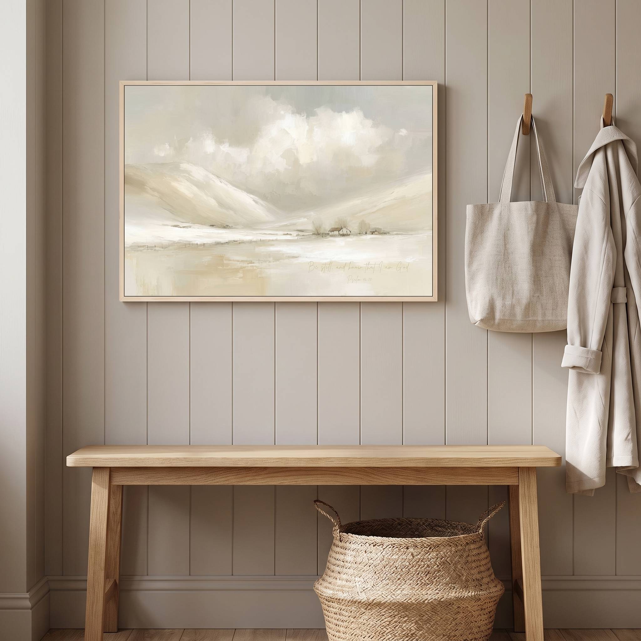Framed vintage Christian winter landscape wall art titled Be Still and Know with Psalm 46:10 verse, displayed on a paneled wall above a light wood bench with wall hooks in a styled entryway.