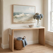 Framed vintage Psalm 46:10 Christian landscape canvas wall art featuring coastal beach ocean waves hanging on a beige wall above a light wood console table with blue hydrangeas and a woven basket.