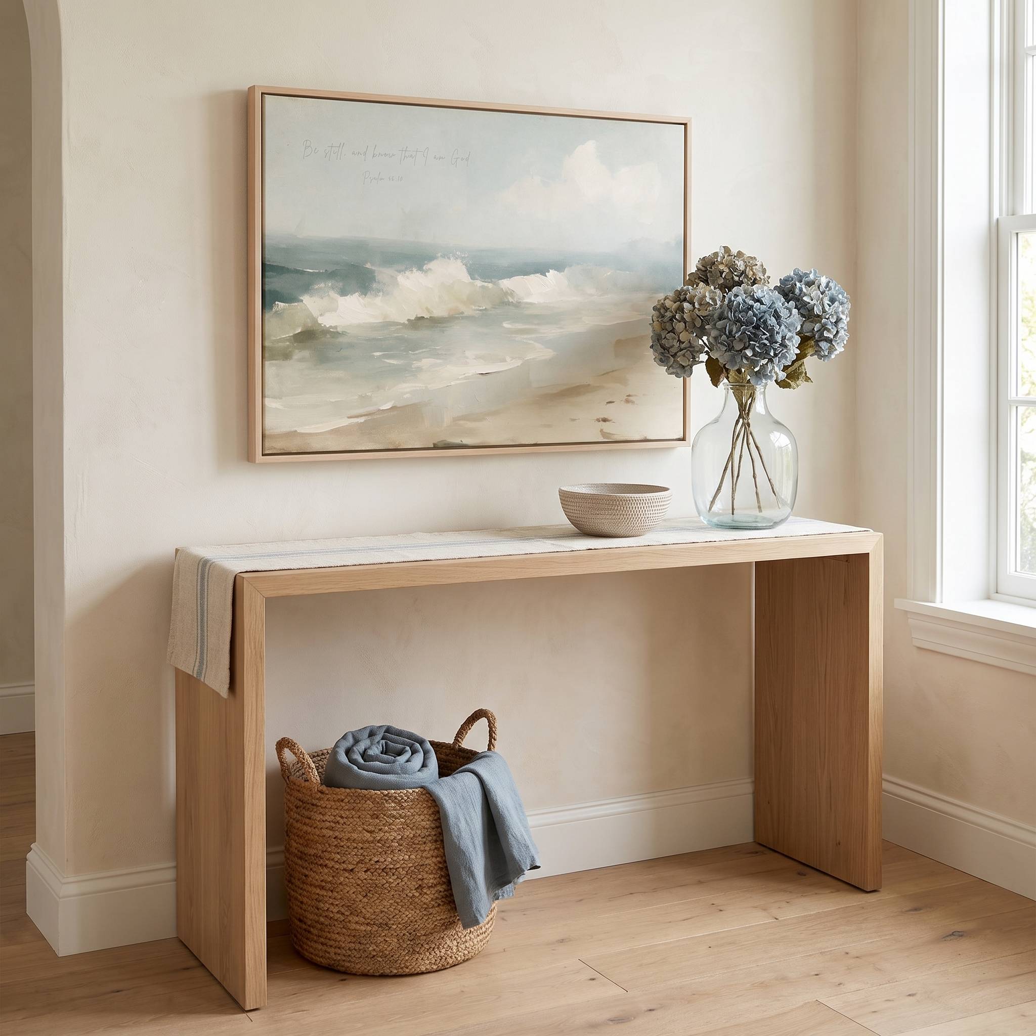 Framed vintage Psalm 46:10 Christian landscape canvas wall art featuring coastal beach ocean waves hanging on a beige wall above a light wood console table with blue hydrangeas and a woven basket.