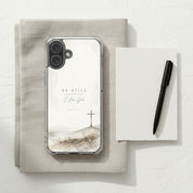 Be Still Psalm 46:10 MagSafe® clear iPhone case featuring soft neutral watercolor landscape artwork with a minimalist Christian cross displayed beside a linen notebook and black pen in a calm modern devotional workspace setting