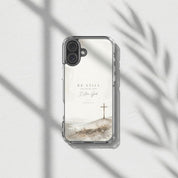 Be Still Psalm 46:10 MagSafe® clear iPhone case featuring soft neutral watercolor landscape artwork with a minimalist Christian cross displayed in warm sunlight with botanical shadows in a peaceful modern devotional aesthetic setting