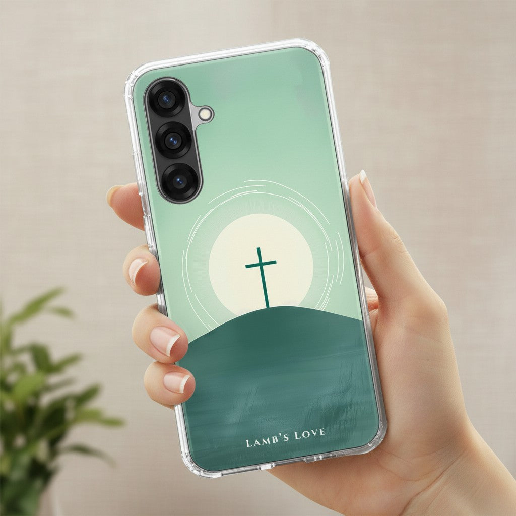 A hand holding a clear-edged Samsung Galaxy phone case featuring a minimalist design of a cross on a hill at sunrise against a soft green background