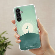 A hand holding a clear-edged Samsung Galaxy phone case featuring a minimalist design of a cross on a hill at sunrise against a soft green background