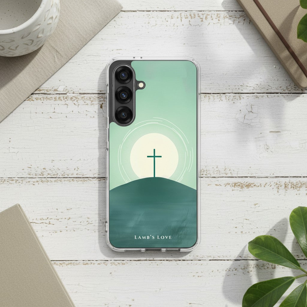 Minimalist Christian Samsung Galaxy case with a cross on a hill at sunrise, displayed on a white painted wooden surface with a notebook and green leaves