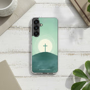 Minimalist Christian Samsung Galaxy case with a cross on a hill at sunrise, displayed on a white painted wooden surface with a notebook and green leaves