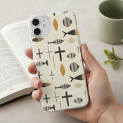 Hand holding a minimalist Christian MagSafe iPhone case featuring a black and gold Ichthys and Cross pattern over an open Bible.