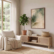 Christian palm leaf wall art displayed above a woven wood console in a light-filled neutral sunroom with a cream armchair and green plant.