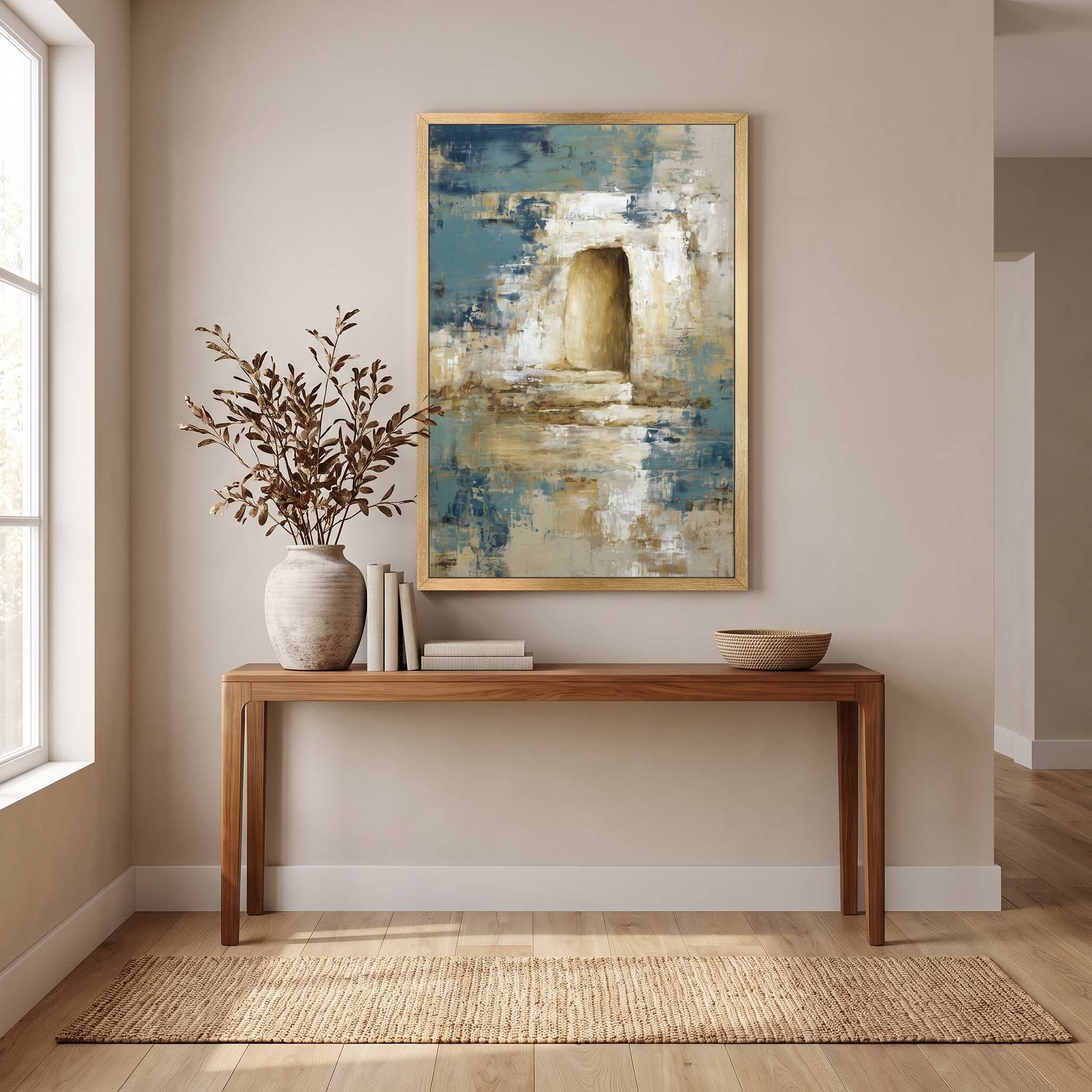 Contemporary Christian abstract empty tomb framed canvas wall art featuring a glowing open tomb motif in blue, beige, and gold displayed on a beige wall above a wooden console table in an organic modern entryway.