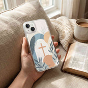 Hand holding a phone case with a cross design, surrounded by a cup of coffee and an open book on a couch.