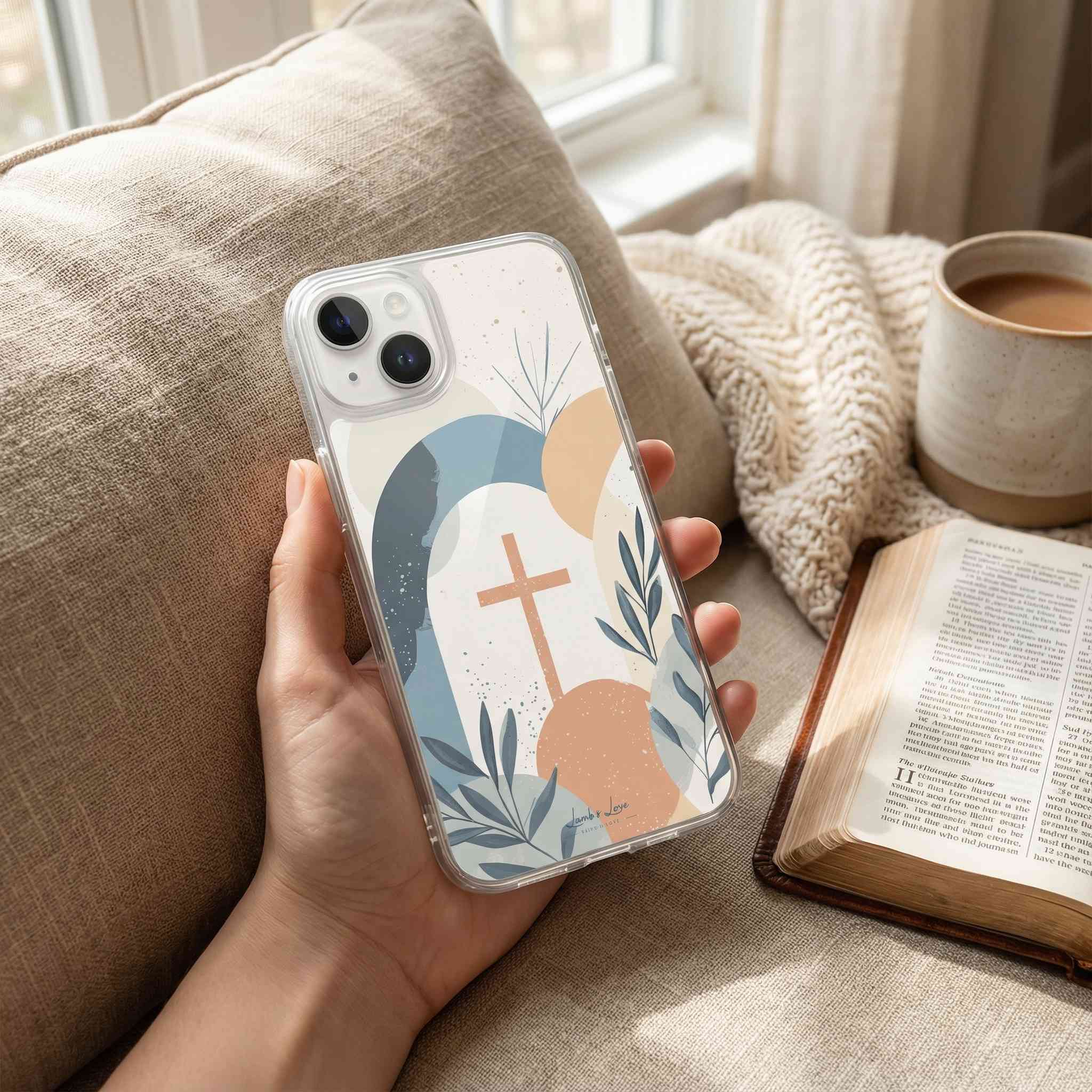 Hand holding a phone case with a cross design, surrounded by a cup of coffee and an open book on a couch.