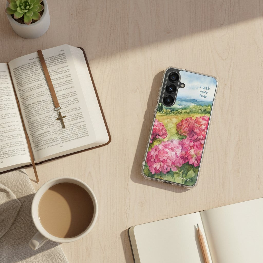 Faith Over Fear Christian Samsung phone case with pink floral landscape design, placed on a light wood desk next to an open Bible with a cross bookmark, a white coffee mug, and a small succulent.