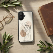 Good Shepherd Christian MagSafe iPhone case beside Bible and eyeglasses on wooden desk with olive branches featuring minimalist artwork of Jesus holding a lamb and warm devotional aesthetic