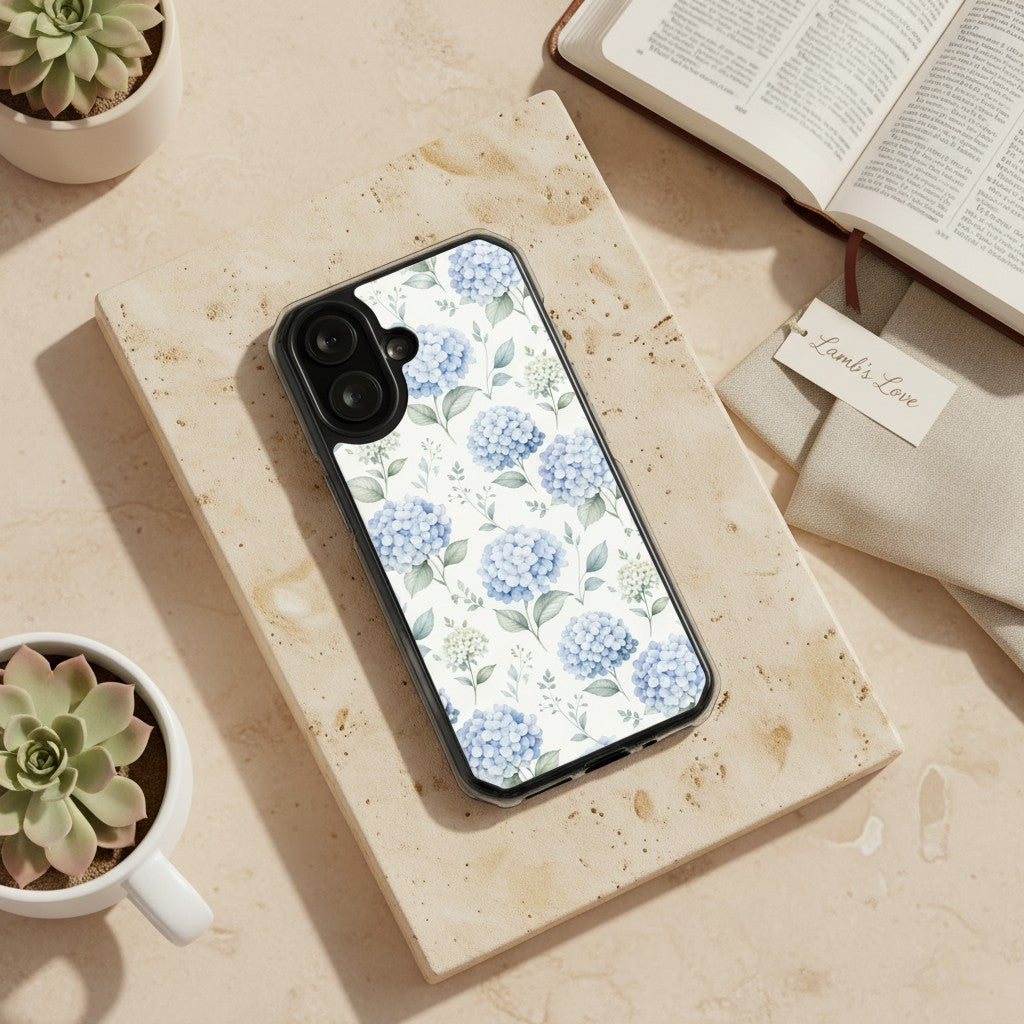 Grace in Bloom MagSafe® clear iPhone case featuring soft blue hydrangea floral artwork displayed beside an open Bible, linen fabric, and ceramic planters in a warm devotional lifestyle setting