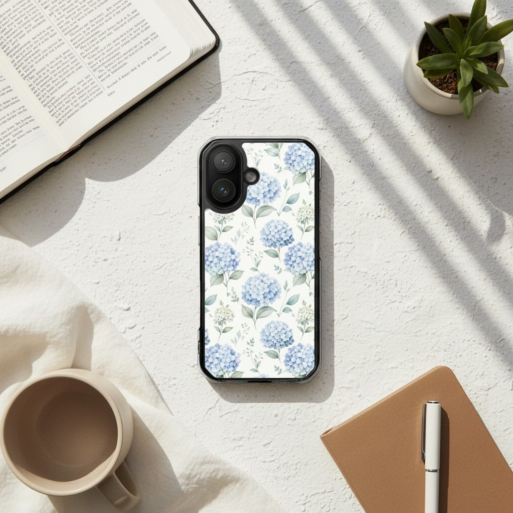 Grace in Bloom MagSafe® clear iPhone case featuring soft blue hydrangea floral artwork displayed on a sunlit desk beside an open book, coffee mug, notebook, and plant in a warm minimalist lifestyle setting