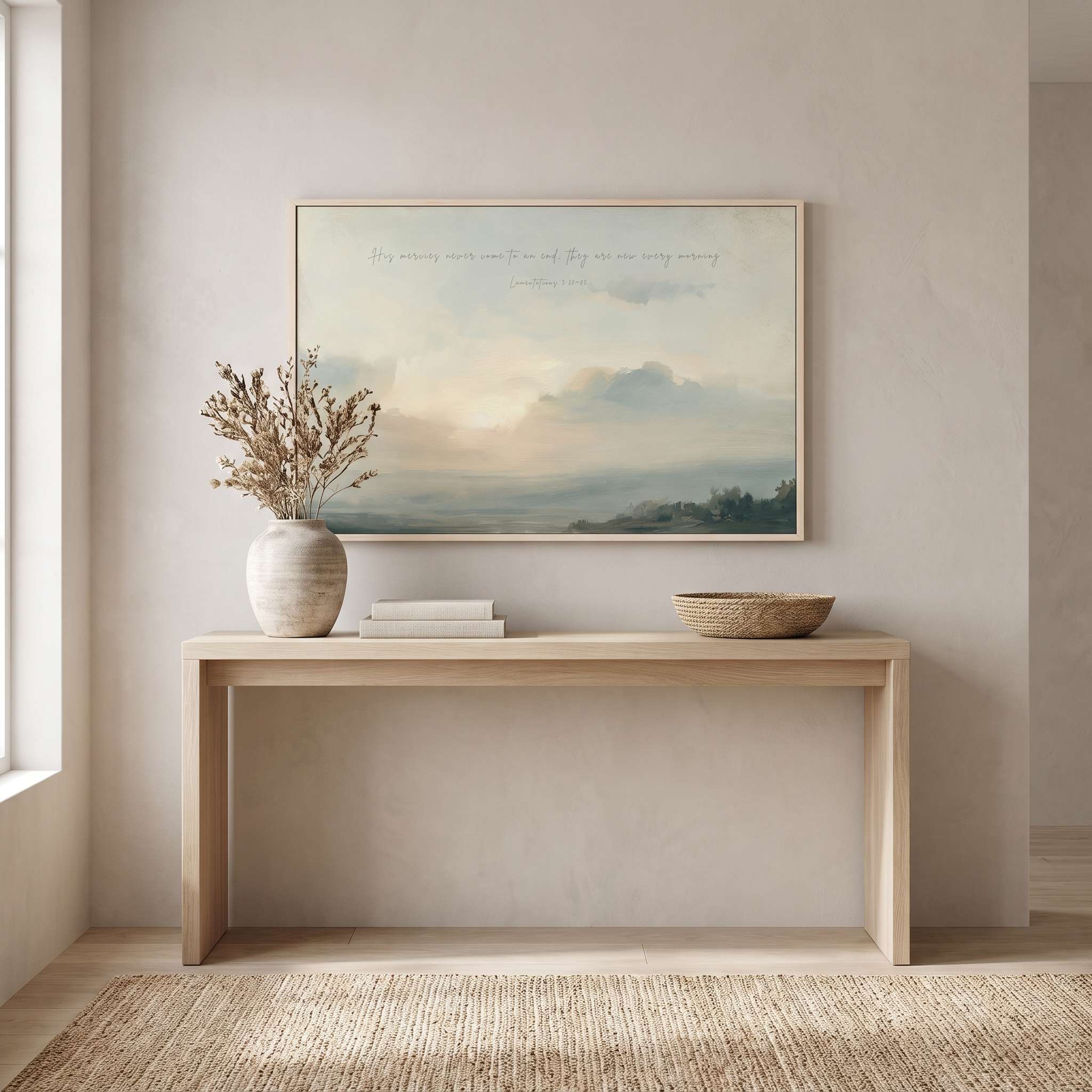Framed vintage Lamentations 3:22-23 Christian landscape canvas wall art featuring a misty sunrise scene hanging above a light wooden console table with stacked books, a vase of dried floral foliage, and a woven bowl in a beige entryway.