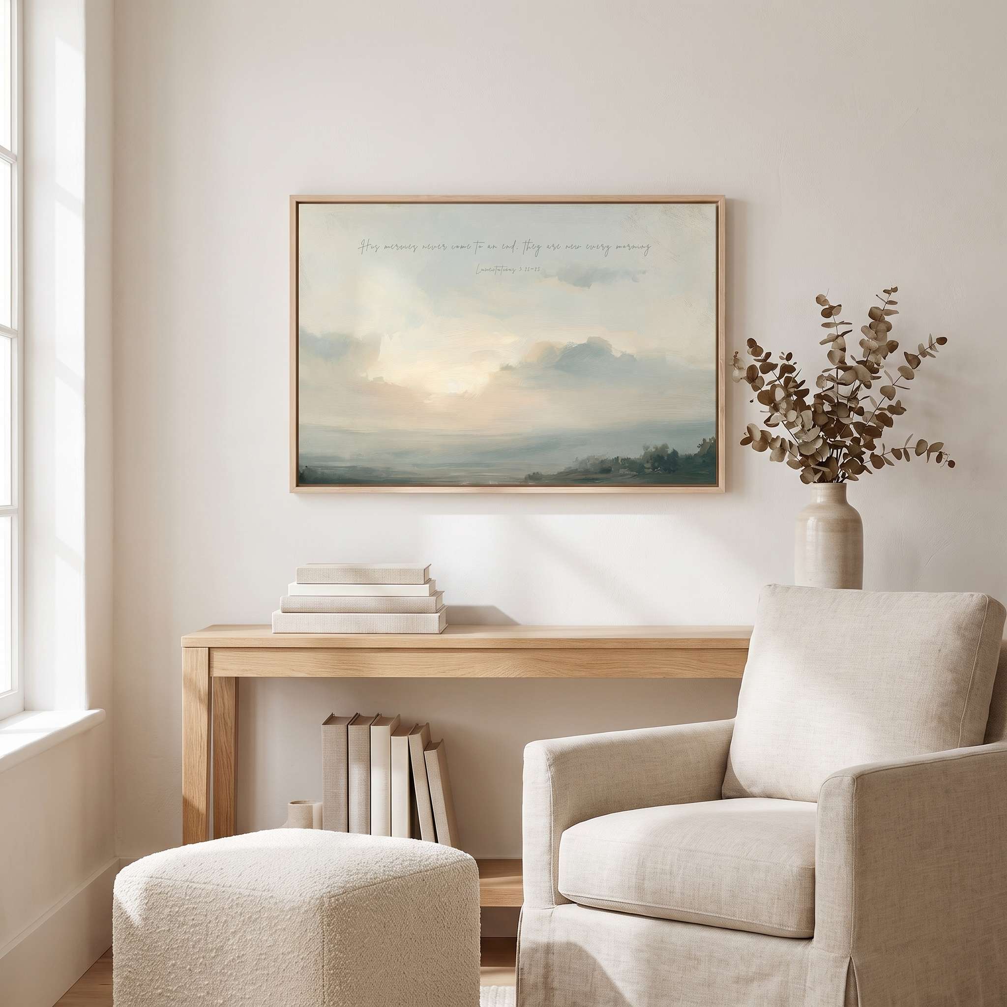 Framed vintage Lamentations 3:22-23 Christian landscape canvas wall art featuring a soft sunrise scene hanging on a light beige wall above a wooden console table, ceramic vase with eucalyptus, and a textured cream armchair with an ottoman in a cozy living room.