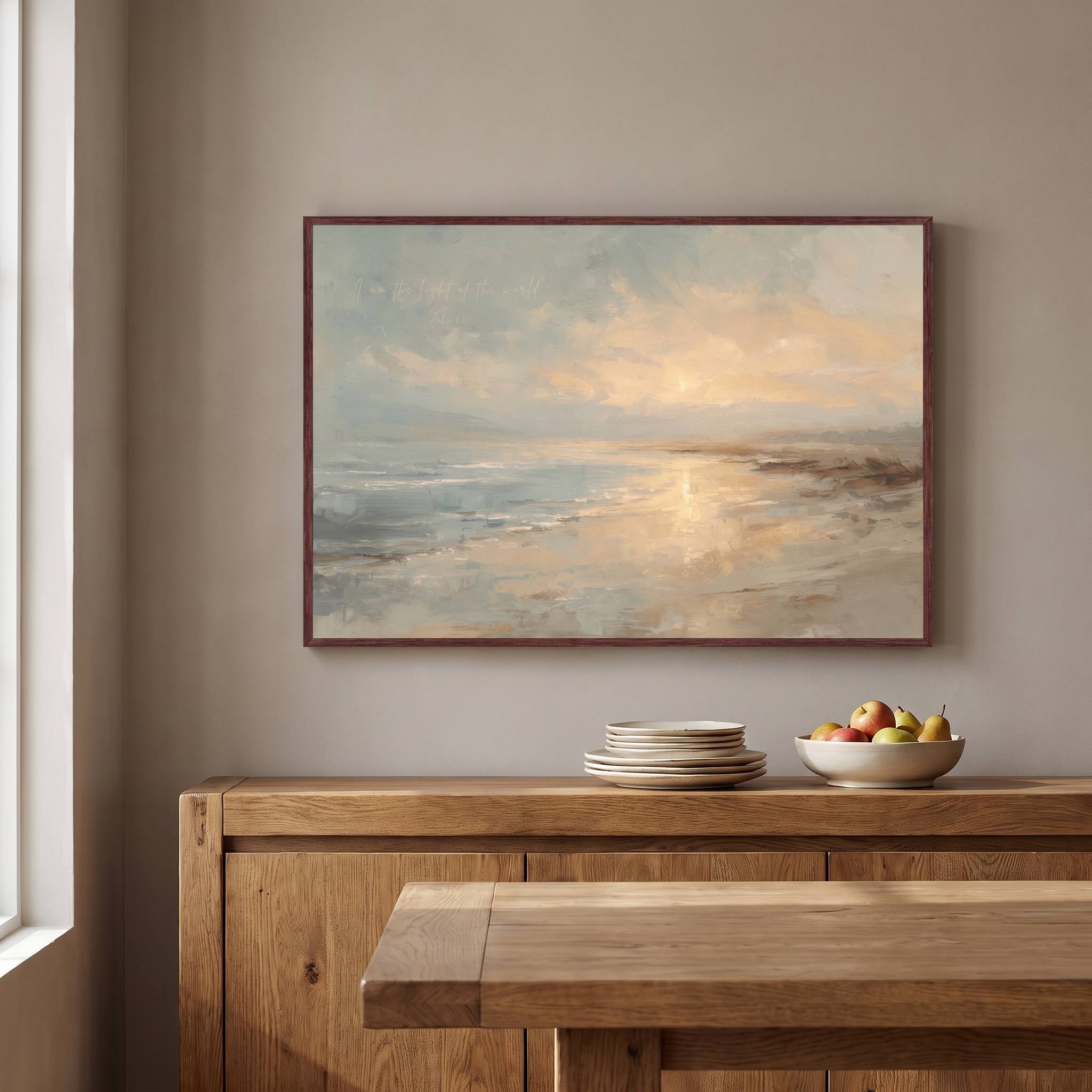 Framed vintage John 8:12 Christian landscape canvas wall art featuring a coastal sunset scene hanging above a wooden sideboard with stacked plates and a fruit bowl in a warm dining room.