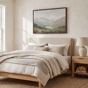 Framed vintage Christian landscape wall art titled I Lift Up My Eyes to the Hills, displaying a peaceful mountain view above a bed in a soft organic modern bedroom.