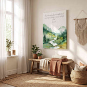 Unframed canvas Christian wall art featuring Isaiah 54:10 Scripture verse with a watercolor forest landscape and river, displayed on a beige wall above a wooden bench with folded blankets in a sunlit room.