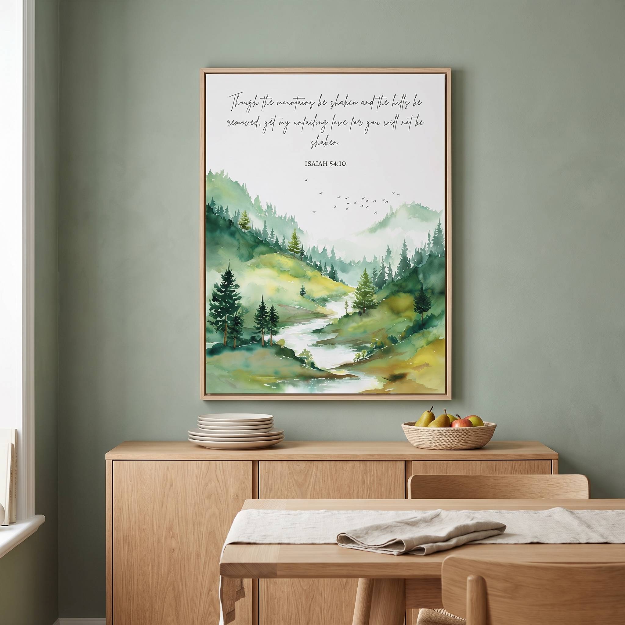 Framed Christian forest landscape wall art titled Isaiah 54:10 featuring evergreen trees, a winding river, and mountains, displayed on a sage green wall above a wooden sideboard in a soft organic modern dining room.