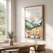 Framed vintage Christian mountain landscape wall art titled Isaiah 6:3 featuring mountains, pine trees, and a field of vibrant wildflowers, displayed on a light wall above a wooden dining table in a bright modern dining room.