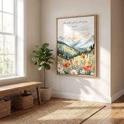 Framed Christian mountain landscape wall art titled Isaiah 6:3 featuring mountains, pine trees, and a field of vibrant wildflowers, displayed on a beige wall above a wooden bench in a bright modern entryway.