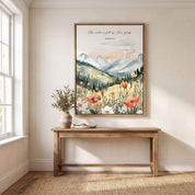 Framed watercolor Christian mountain landscape wall art titled Isaiah 6:3 featuring mountains, pine trees, and a field of vibrant wildflowers, displayed on a beige wall above a wooden console table in a bright modern entryway.