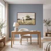 A framed landscape art print hangs on a dusty blue-gray wall in a modern, sunlit home office. The room features a light wood desk, a matching upholstered chair, a cream ceramic table lamp, and neutral-toned decor.