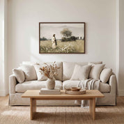 A framed landscape art print hangs on a light beige wall above a cream-colored sofa in a styled living room setting. The print depicts a woman standing in a field of white flowers under a tree.