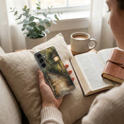 Person holding a Samsung phone with a Christian scenic case, sitting on a couch with an open book and cup of coffee.