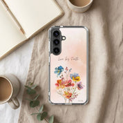 Samsung phone case with floral design and 'Live by Faith' text on a wooden surface with mugs and a notebook.