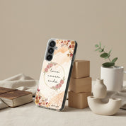 Love Never Ends Christian Samsung Galaxy phone case with a pink floral wreath and 1 Corinthians 13:8 verse, displayed in a lifestyle studio setting with a book and modern ceramic decor.