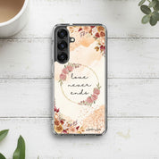 Love Never Ends Christian Samsung Galaxy phone case with a delicate pink floral wreath design and 1 Corinthians 13:8 Bible verse on a white wood background.