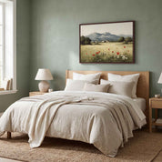 A framed horizontal vintage-style Christian landscape painting depicting a meadow of red wildflowers and mountains inspired by Luke 12:27, displayed above a bed with light beige linens in a modern bedroom.