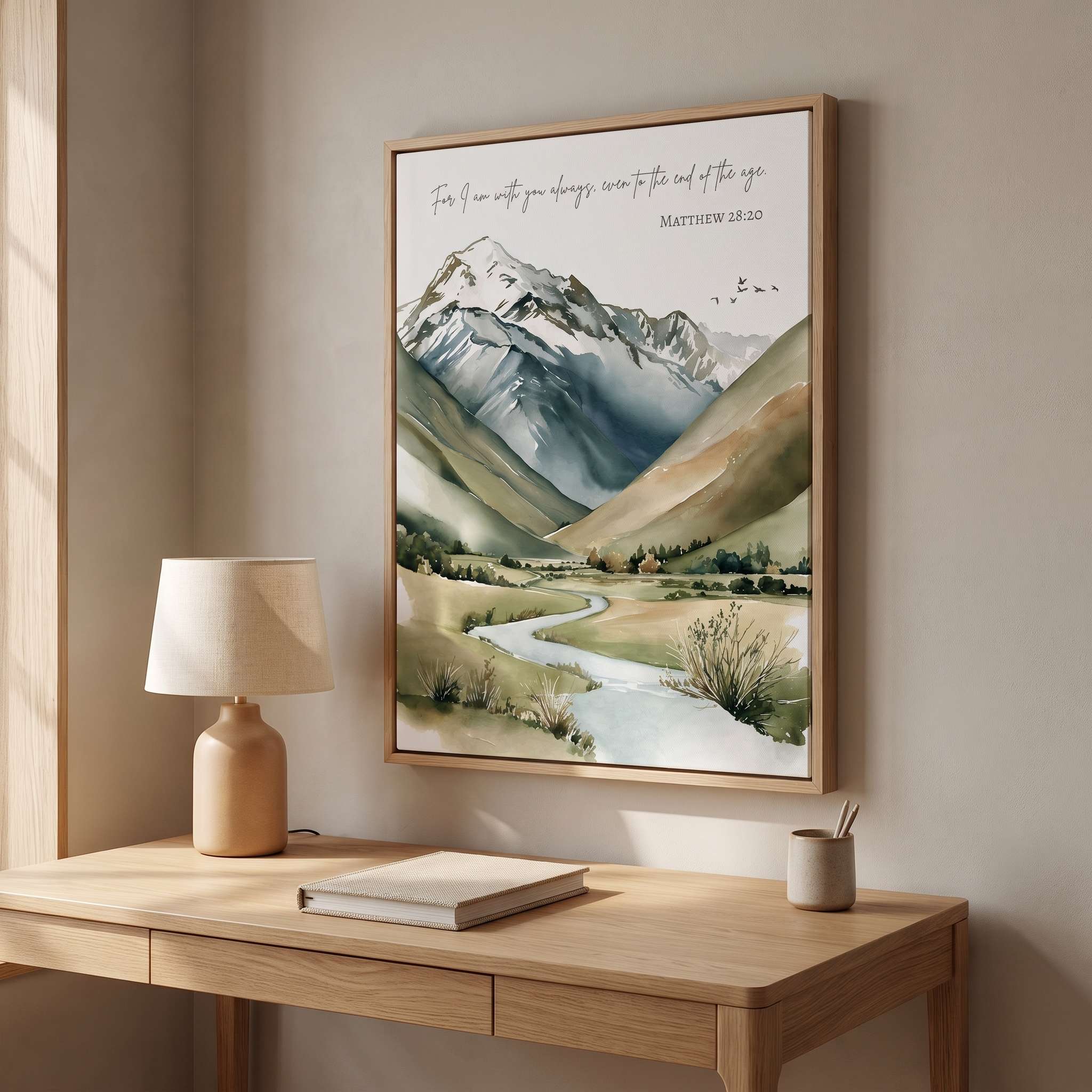 Framed vintage Christian mountain landscape wall art titled Matthew 28:20 featuring mountains, a winding river, and valley, displayed on a beige wall above a wooden desk with a ceramic lamp in a bright modern home office.