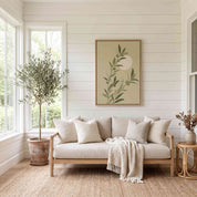 Minimalist olive branch vertical framed canvas Christian wall art in soft sage green displayed on a white shiplap wall above a light beige sofa in an organic modern sunroom.