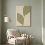 Minimalist olive leaf vertical framed canvas wall art displayed on a sage green wall above a light wood armchair in an organic modern reading nook.