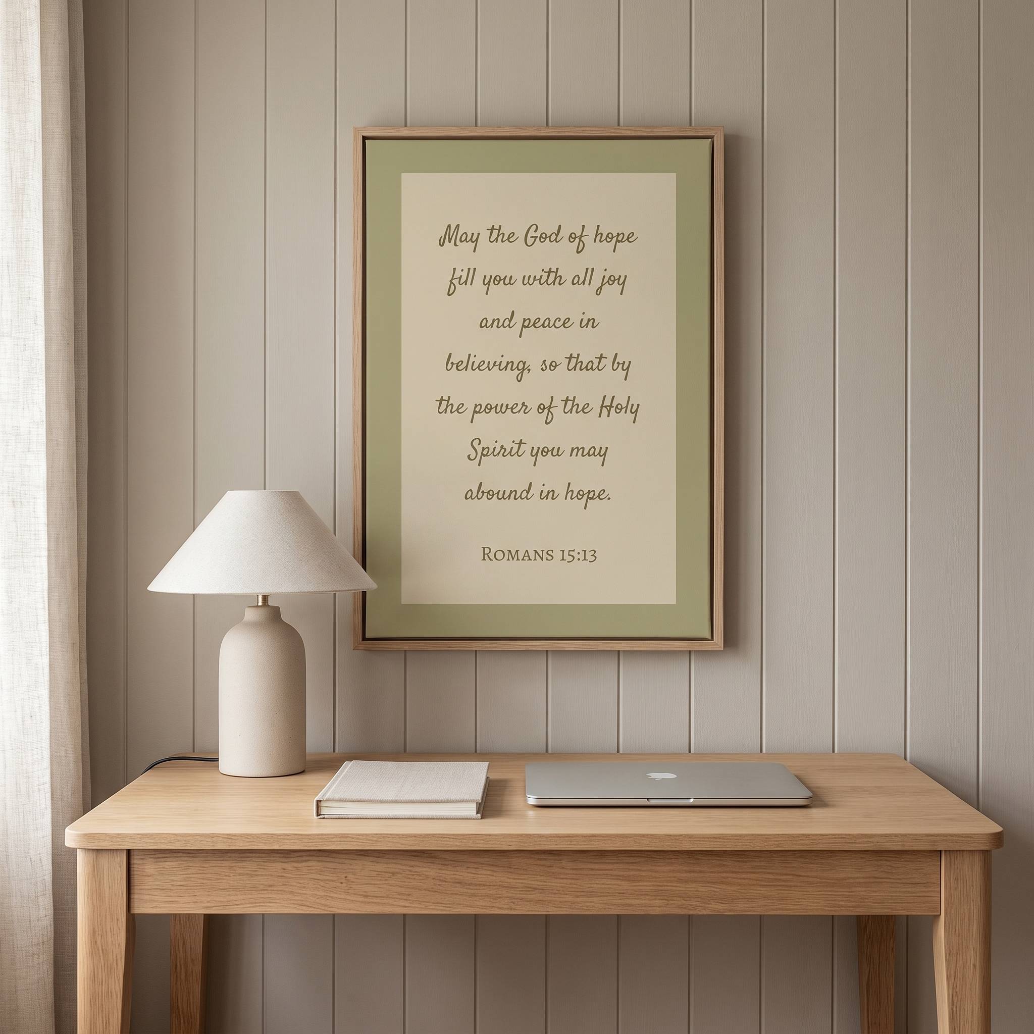 Minimalist Romans 15:13 Scripture vertical framed canvas wall art in soft sage green displayed on a vertical paneled wall above a light wood desk in an organic modern workspace.