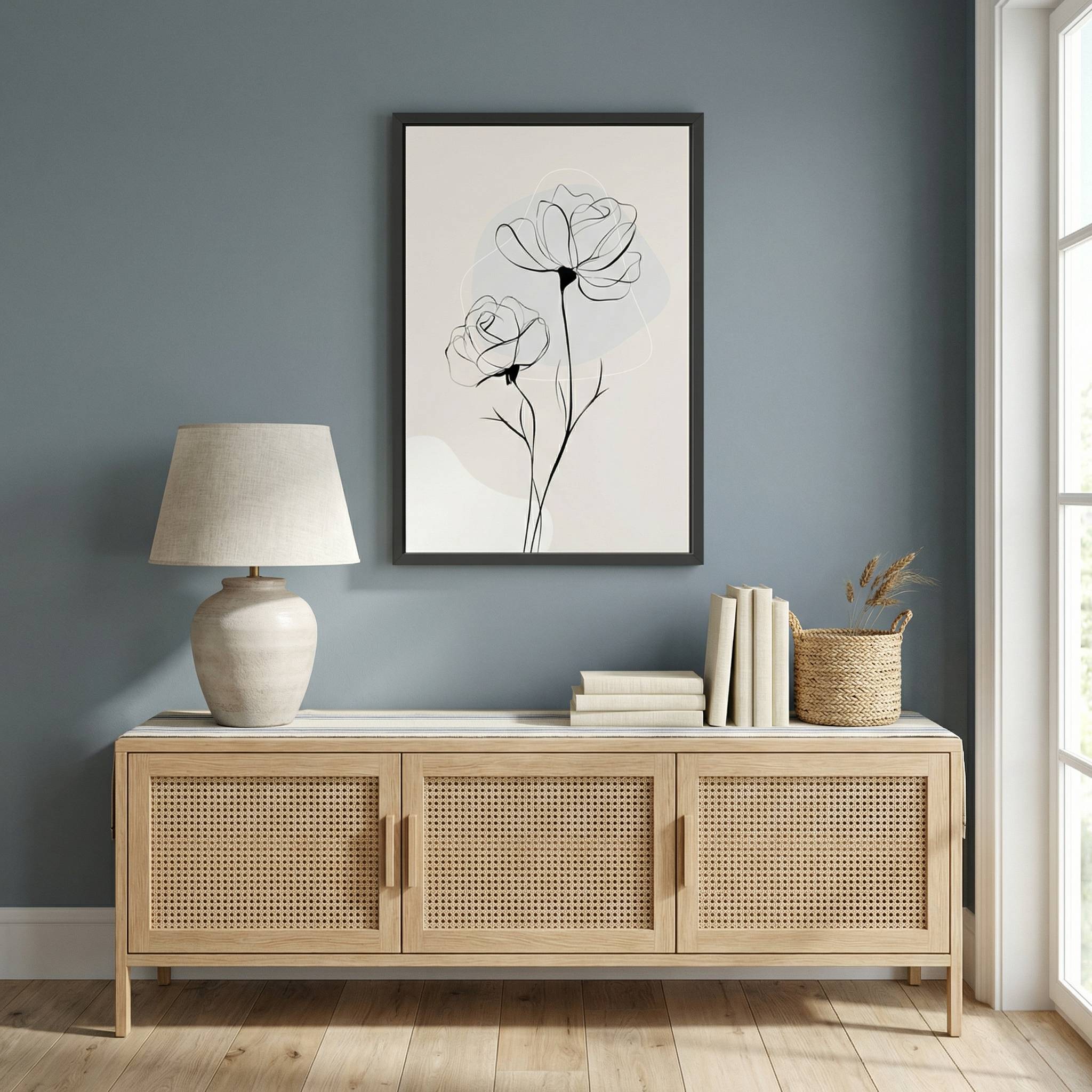 Minimalist Rose Line Art in a black frame, styled on a blue-gray wall above a light wood cane console table with a ceramic lamp and books