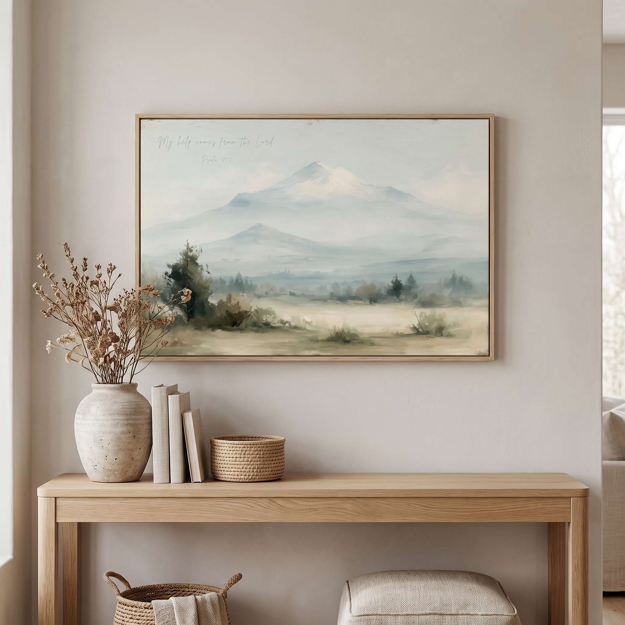 Framed vintage Psalm 121:2 Christian landscape canvas wall art featuring snow-capped mountains hanging above a light wooden console table with stacked books, a ceramic vase with dried flowers, and a woven basket in a beige entryway.