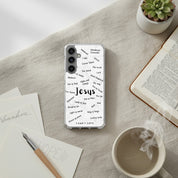 A clear Samsung Galaxy phone case with black typography of the Names of Jesus, placed on a beige textured surface next to an open book, a steaming white coffee mug, a small succulent, and a pen.