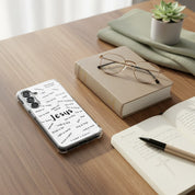 A clear Samsung Galaxy phone case with black biblical names of Jesus typography, resting on a wooden desk next to a pair of glasses, an open Bible with a pen, and a small potted succulent.