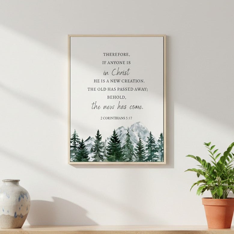 Framed Christian wall art featuring 2 Corinthians 5:17 with watercolor pine trees and mountains, displayed above a wooden desk in natural light.