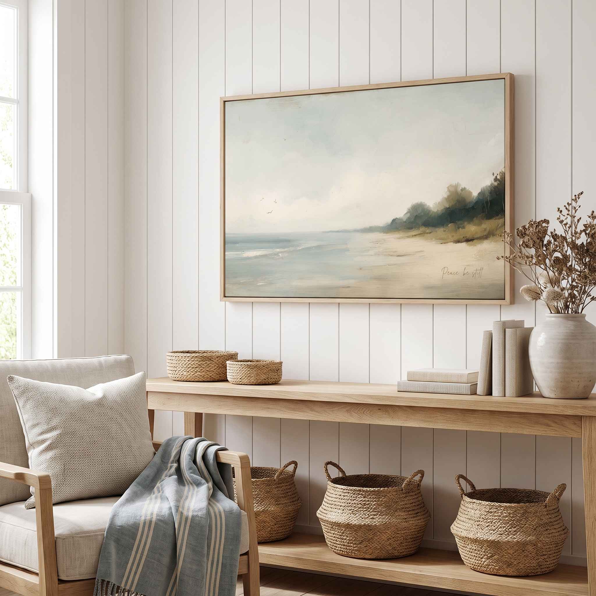 Framed vintage Mark 4:39 Christian landscape canvas wall art featuring a coastal beach scene hanging on a white shiplap wall above a light wooden console table with woven baskets and an armchair in a cozy living room.