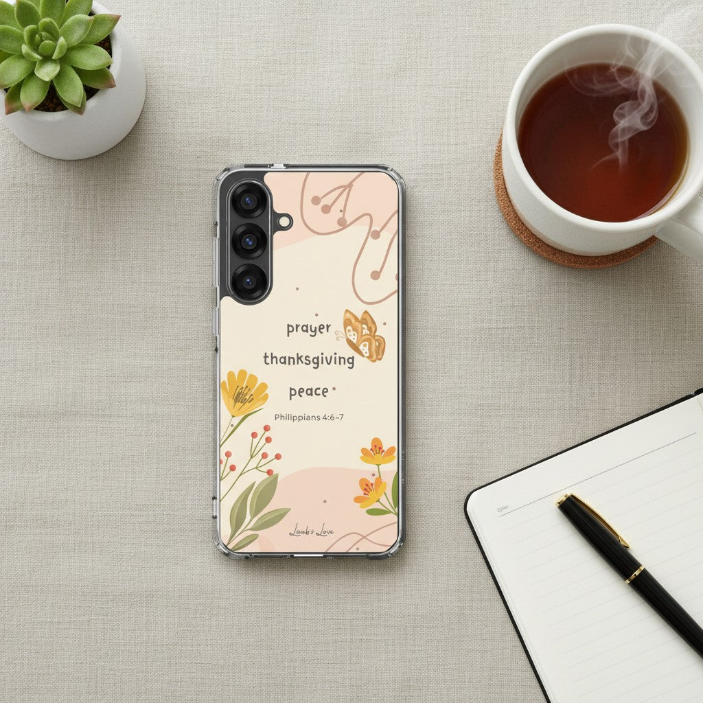 Prayer Thanksgiving Peace Christian Samsung phone case with warm floral design and Philippians 4:6-7 Bible verse, pictured in a flat lay on a beige surface next to a steaming cup of tea, a notebook, and a black pen.